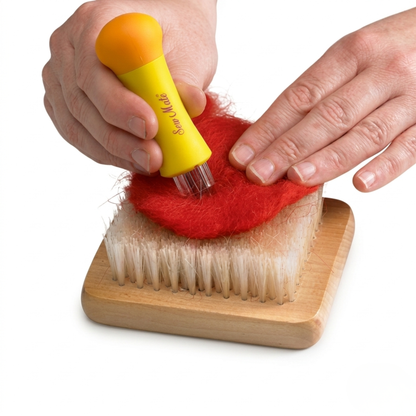 Hands using a yellow multi-needle pen tool to sculpt red wool fiber on a square wooden needle felting brush mat.