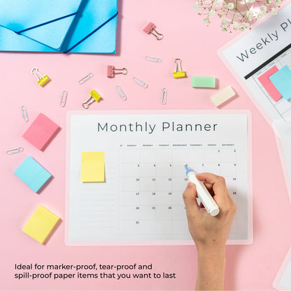 Hand marking a monthly planner sheet with a dry erase pen over a bright pink background setup.