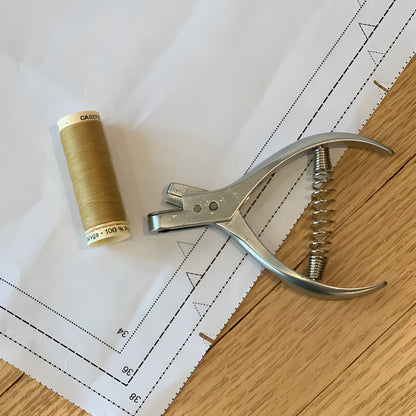 Professional pattern notcher pliers resting on white blueprint design paper next to a spool of gold sewing thread.