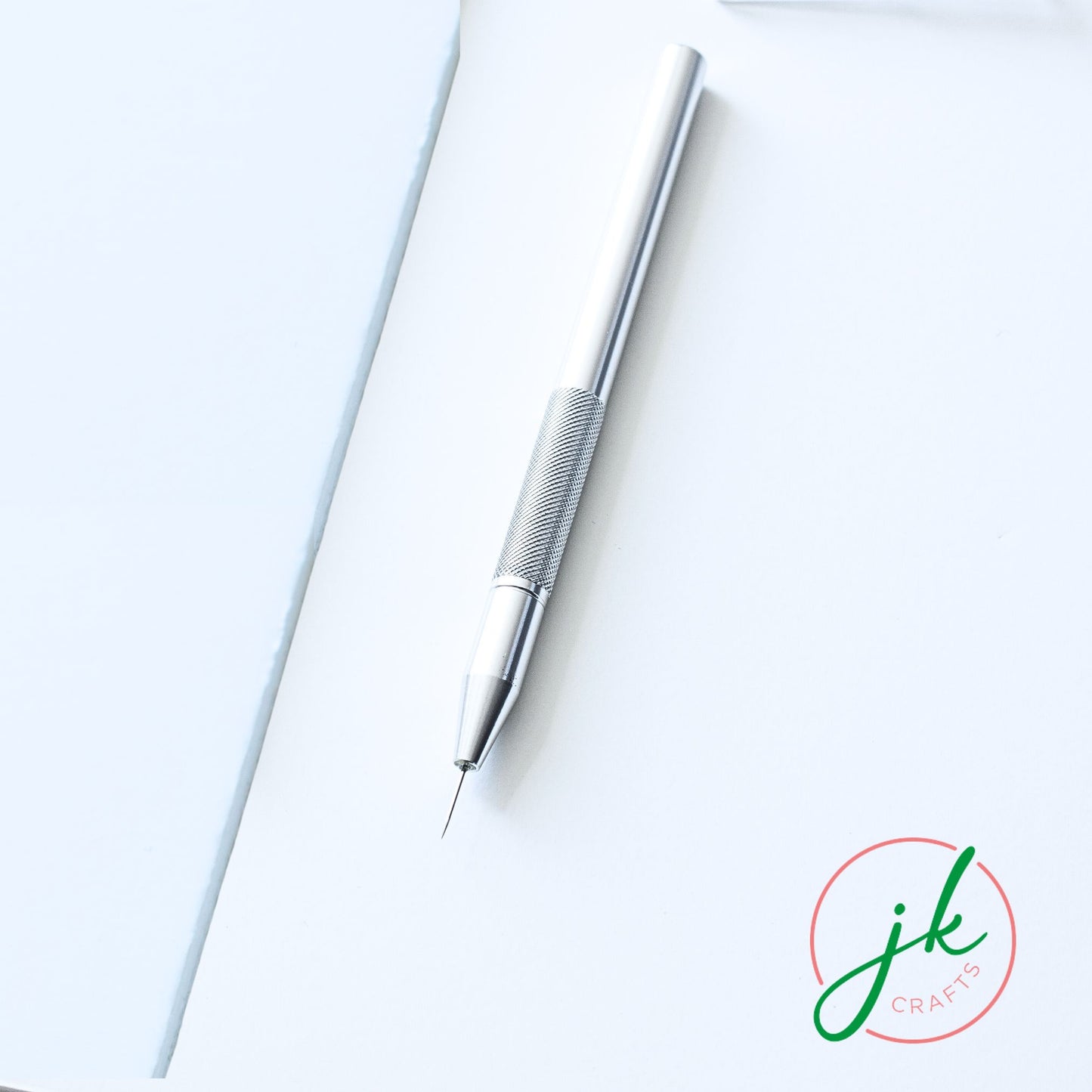 Sleek solid aluminium single-pin perforating pen resting diagonally on a crisp white workspace background.