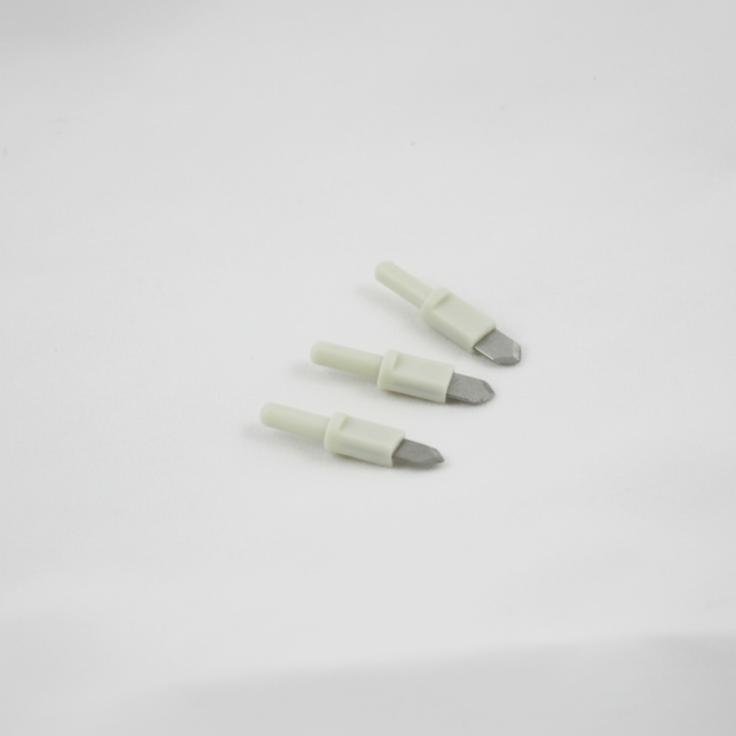 Angled view of three small white and silver graphic art utility trimmer blades resting on a clean white desk surface.